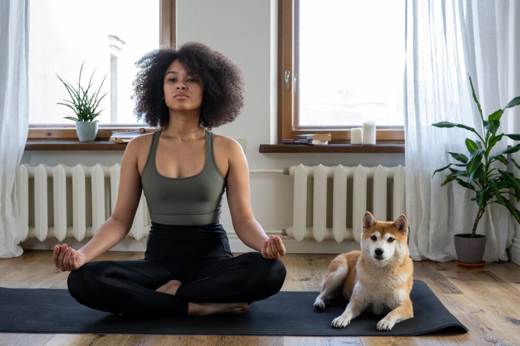 A woman sits beside her dog while meditating on a yoga mat.