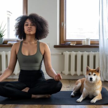 A woman sits beside her dog while meditating on a yoga mat.